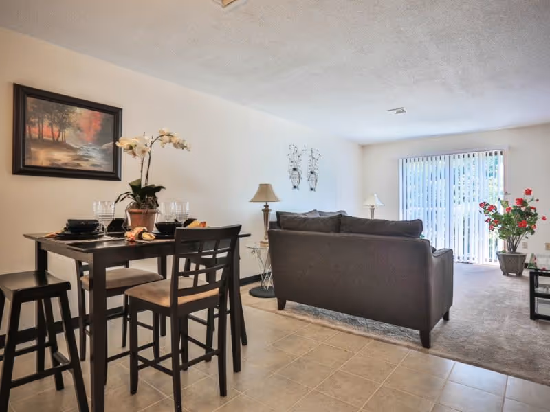 A bright living and dining area in a senior living facility. The dining area features a dark wooden table set with four chairs and place settings, including plates, bowls, and glasses. A potted orchid plant is centered on the table. The living area has a dark sofa facing away from the dining table, with two lamps on side tables and a large window with vertical blinds letting in natural light. A painting hangs on the wall above the dining table, and a potted plant is near the window.