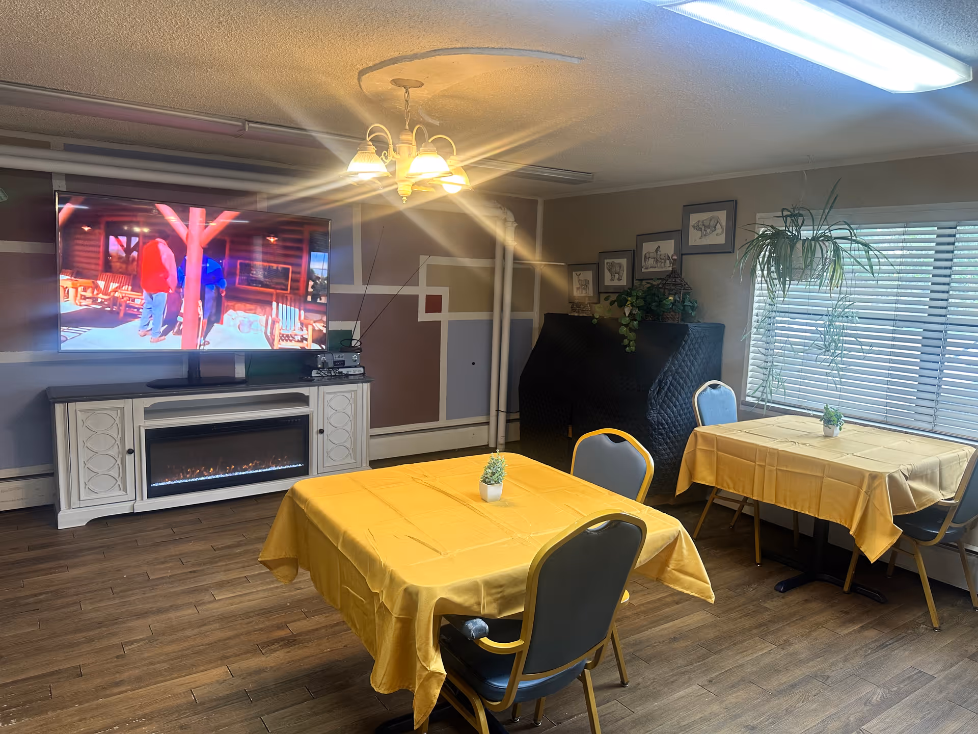 A cozy common area with two tables covered in yellow tablecloths, each with a small potted plant. There are several chairs around the tables. A large flat-screen TV is mounted on the wall above a white electric fireplace. The walls have a geometric design, and there is a hanging light fixture on the ceiling. A window with blinds and a hanging plant is on the right side of the room.