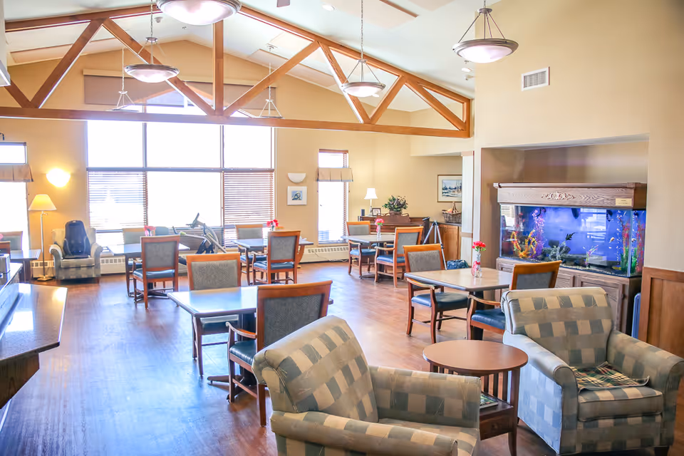 Bright senior living lounge with tables, chairs, and a large aquarium.