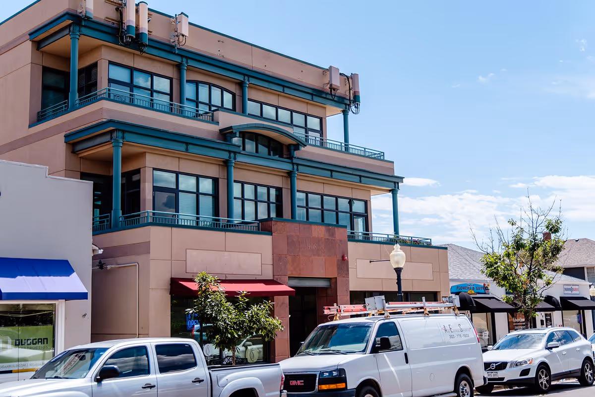 Three-story beige building with teal accents, glass windows, and parked vehicles along street.