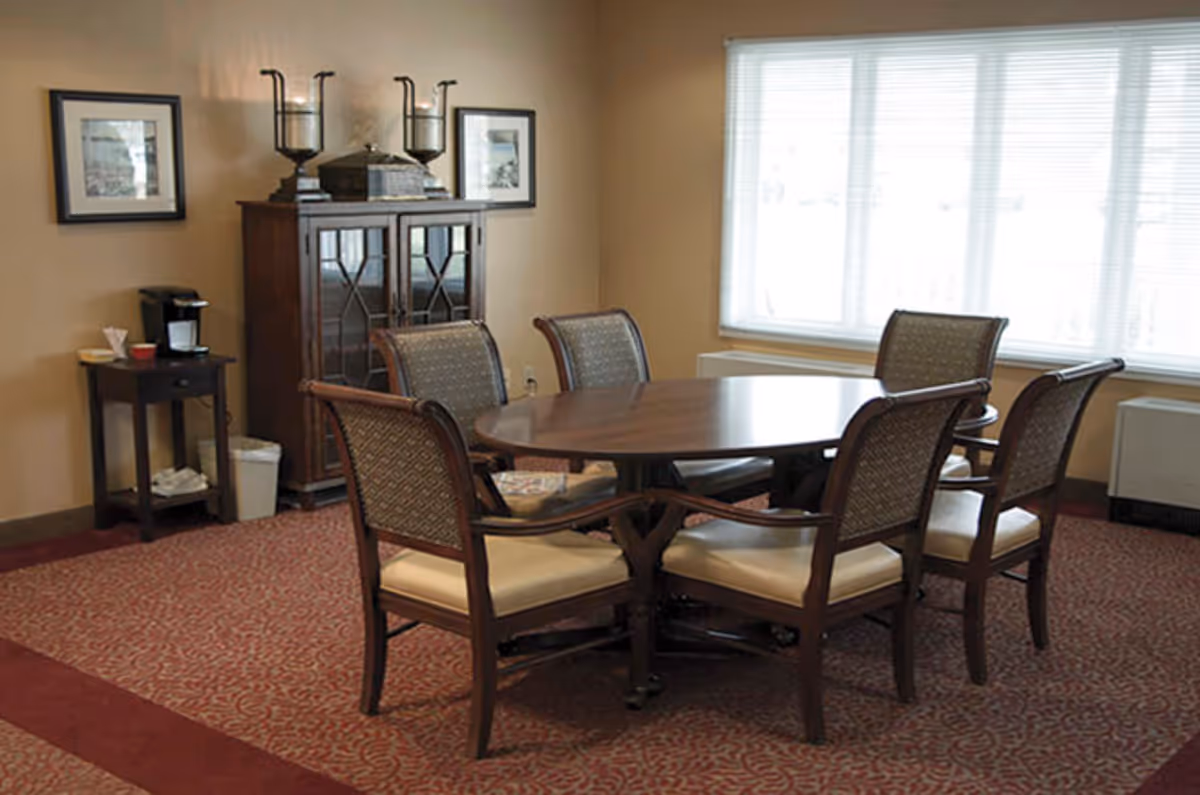 Round wooden table with six upholstered chairs in a carpeted dining/meeting room with a cabinet and coffee station by a large window.