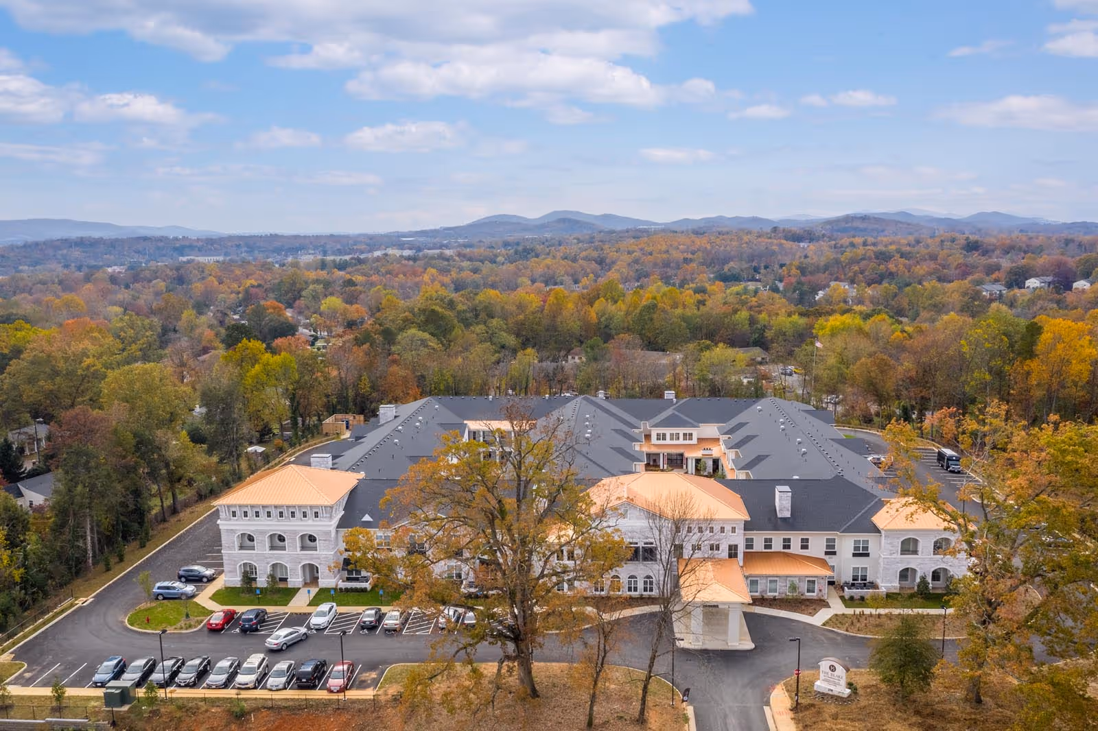 A large senior living facility with gray roof, orange accents, and autumn trees.