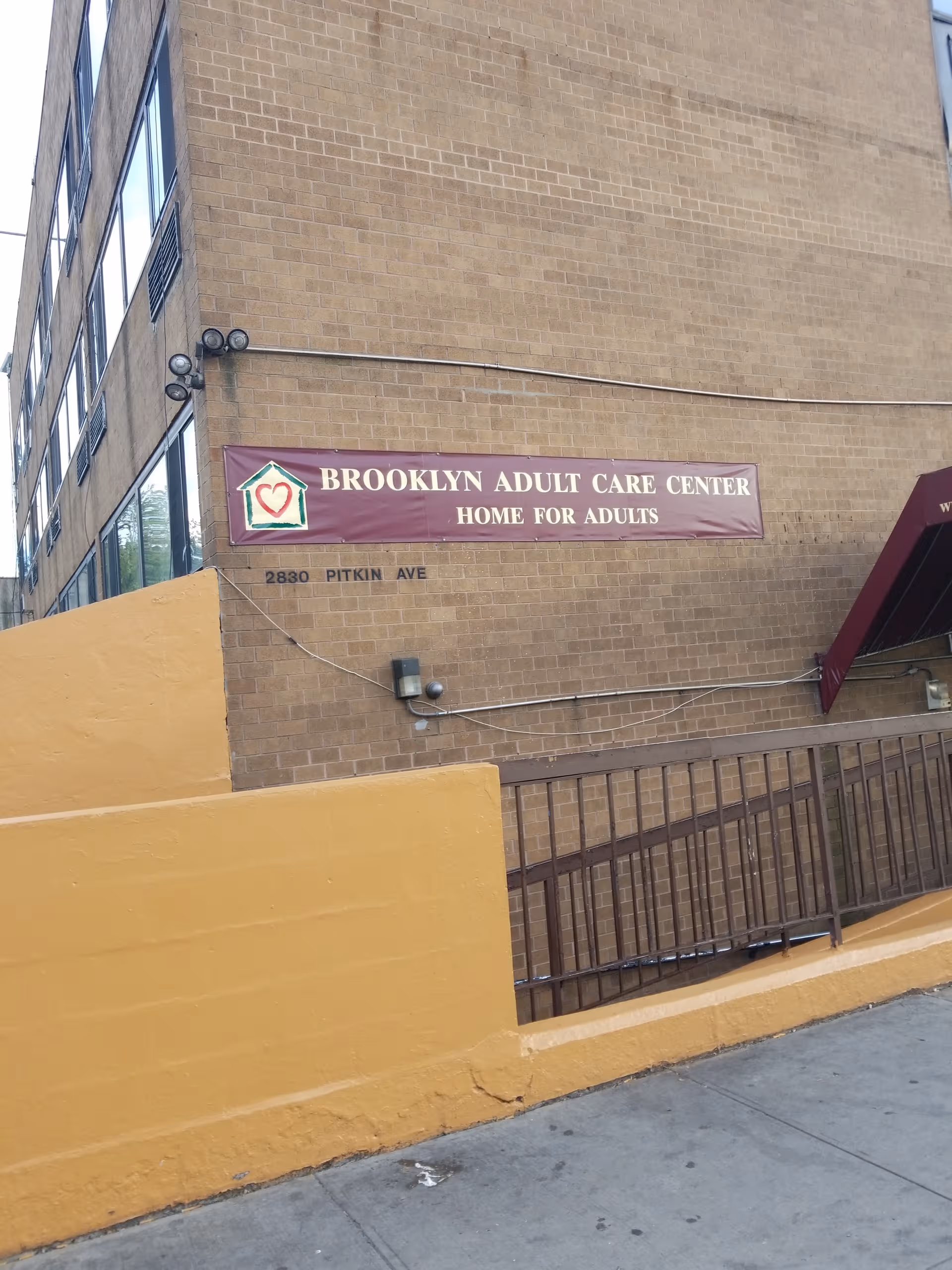 Brick building with Brooklyn Adult Care Center sign and yellow concrete barrier at street.