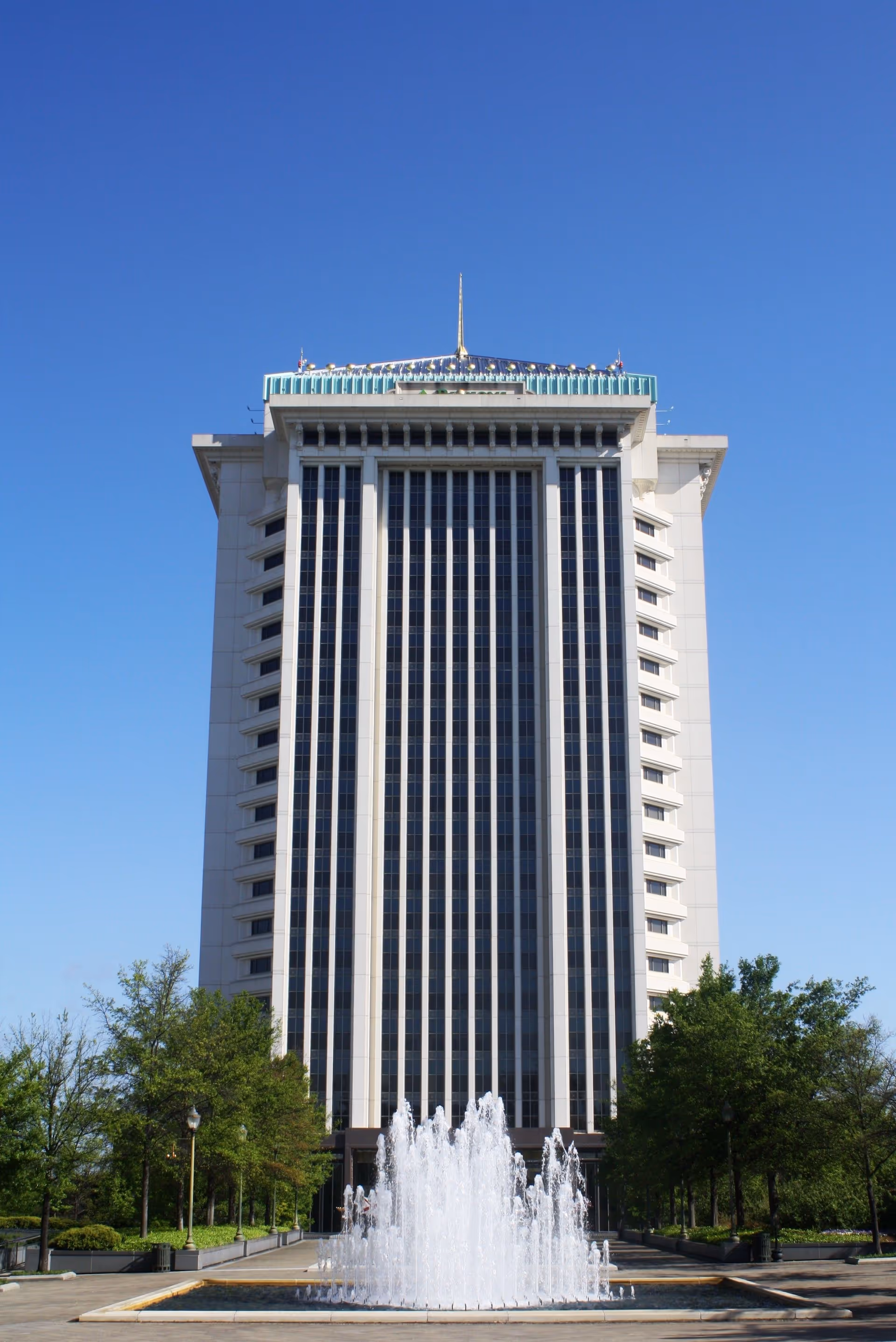 Tall modern high-rise building with a glass façade framed by trees and a fountain.