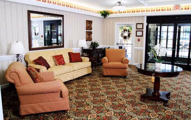 A cozy senior living room with beige sofa, striped chairs, lamps, coffee table, and patterned carpet.