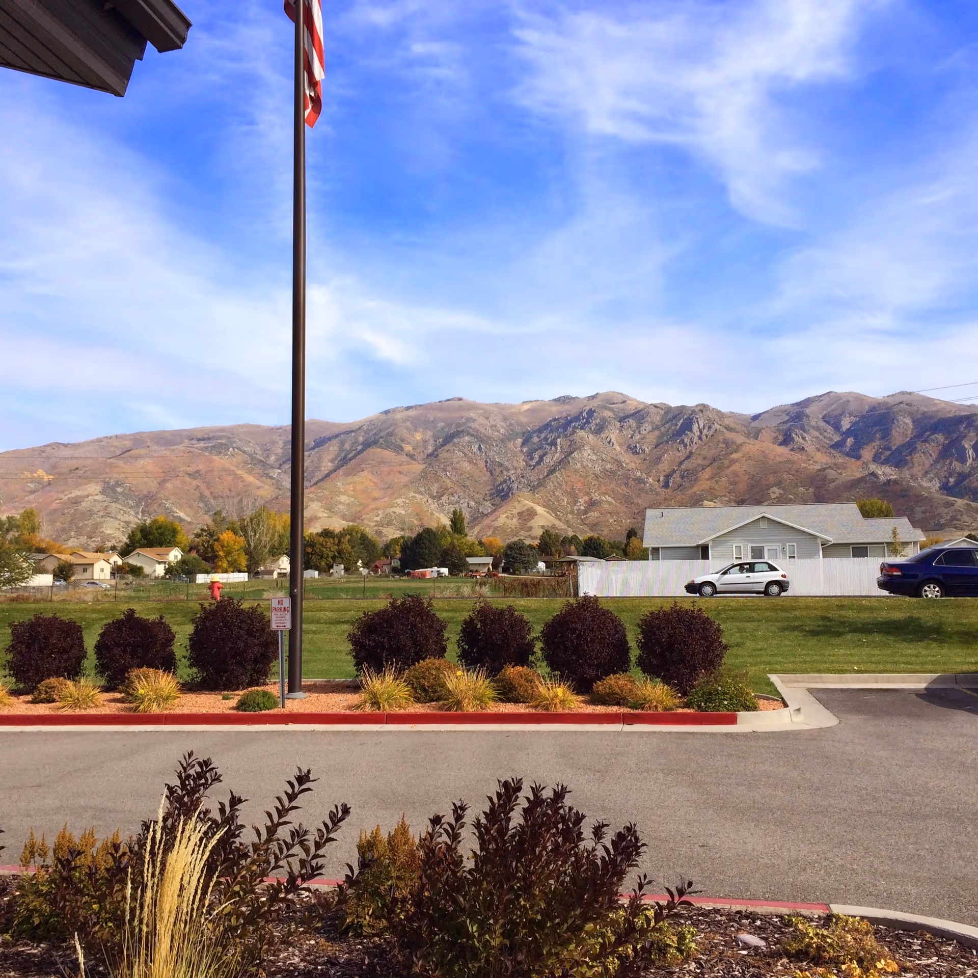 View of a landscaped parking area with bushes and an American flag on a tall flagpole. In the background, there are houses, a white fence, parked cars, and a range of mountains under a partly cloudy blue sky.