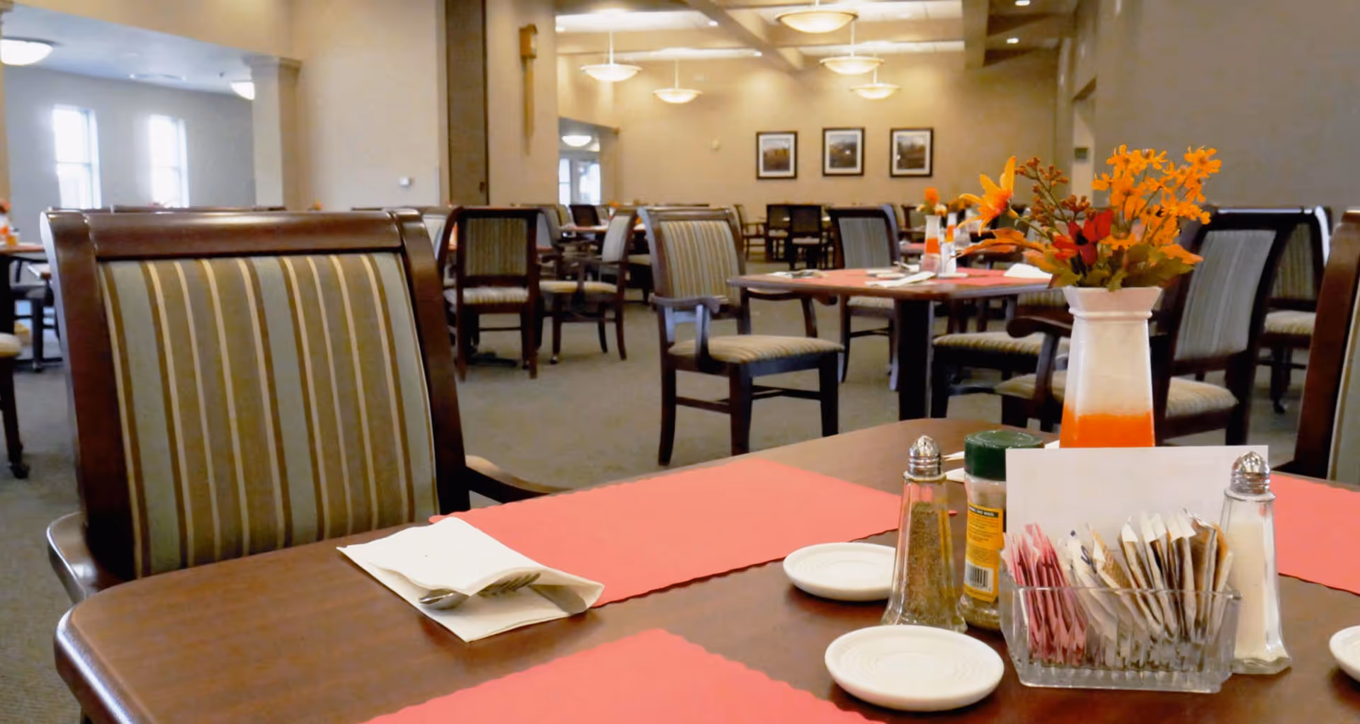 Spacious dining room with tables and striped chairs set with red placemats, condiments, and a vase of orange flowers.