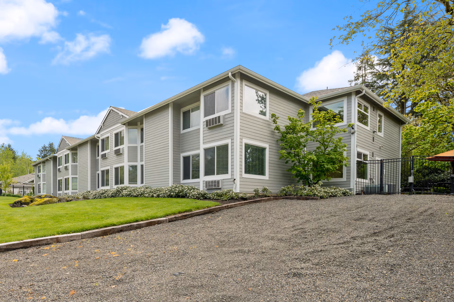 Exterior view of a two-story senior living facility building with light gray siding, multiple windows, and a well-maintained lawn with shrubs and trees under a blue sky with some clouds.
