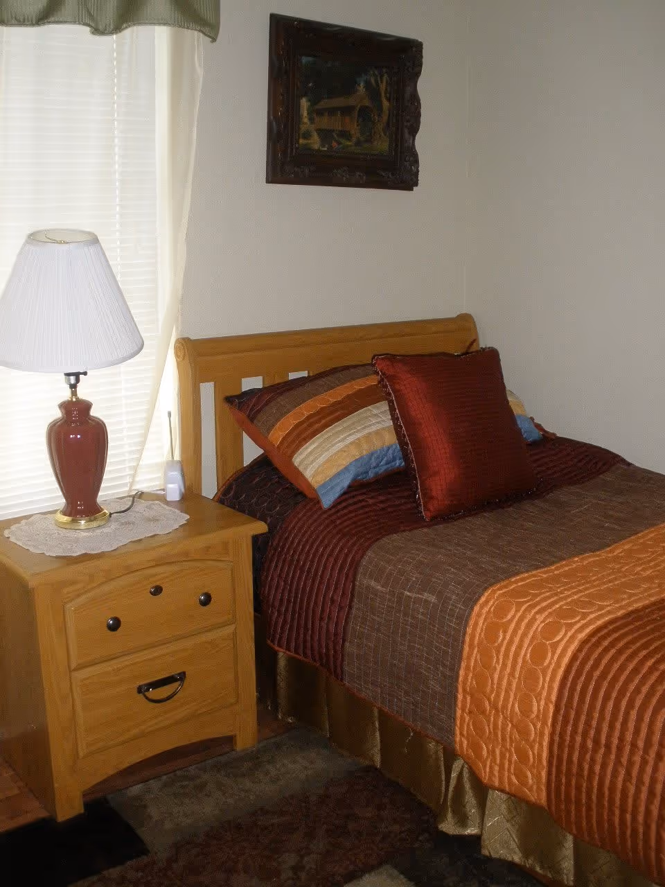 A cozy bedroom corner featuring a wooden bed with a multi-colored quilt and pillows in shades of brown, orange, and red. Next to the bed is a wooden nightstand with two drawers, a red table lamp with a white lampshade, and a small white device. A window with white blinds and light curtains is behind the nightstand, and a framed painting hangs on the wall above the bed.