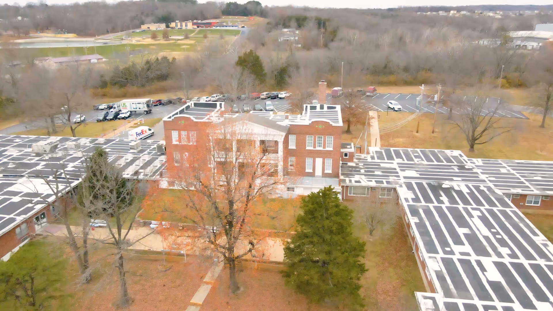 Aerial view of a large brick building complex with multiple wings and a central taller section, surrounded by trees and parking lots with several vehicles including a FedEx truck. The landscape appears to be in late autumn or winter with leafless trees and brown grass.