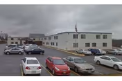 Exterior view of a light-gray building with a parking lot and several cars.