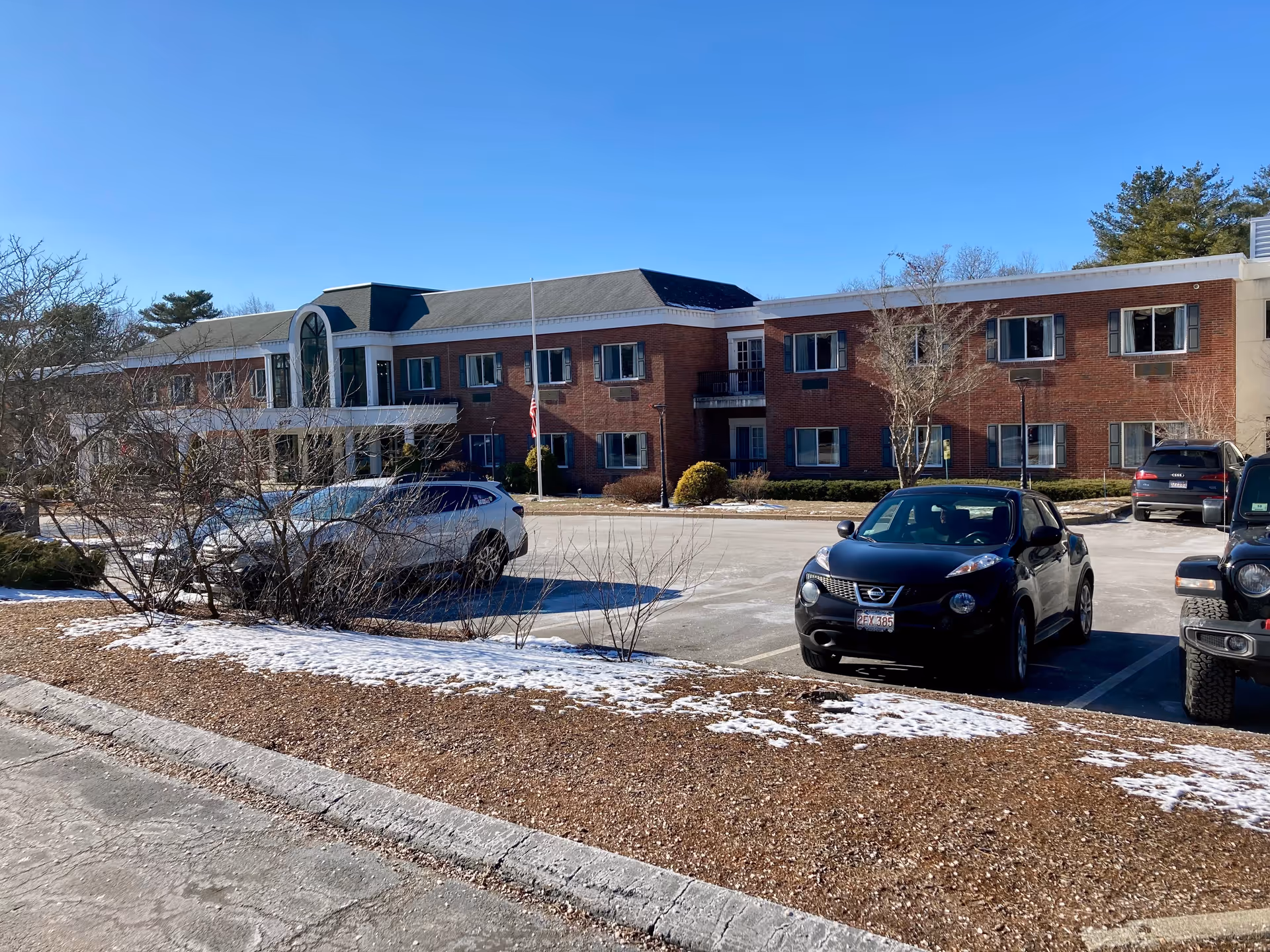 Two-story brick senior living building with a parking lot, parked cars, and light snow on landscaping under a clear blue sky.