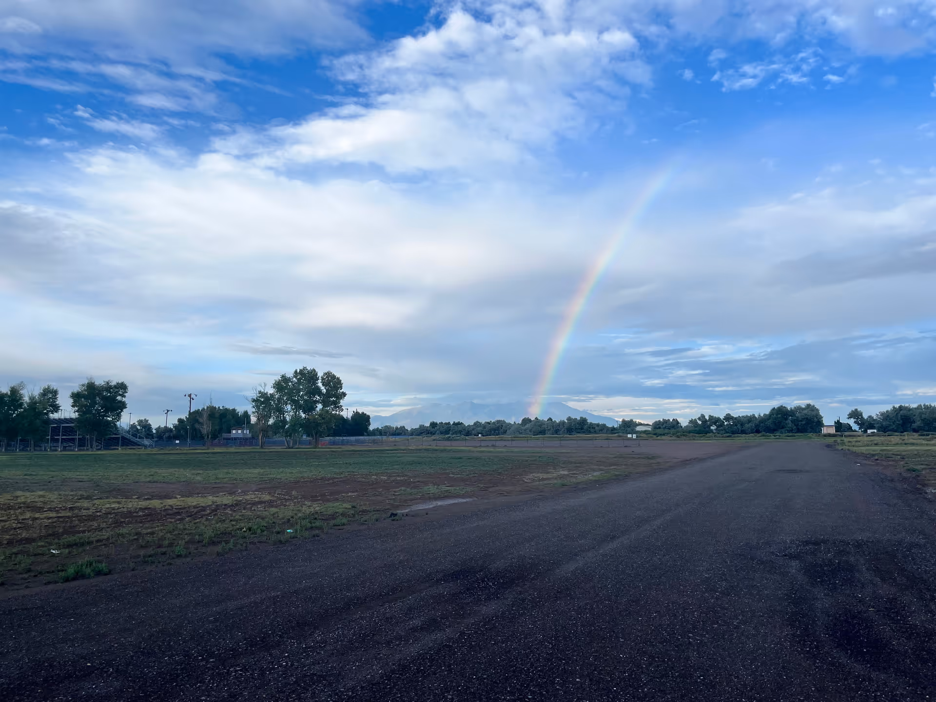 Open field with a dirt road, trees, and a rainbow under a cloudy blue sky.