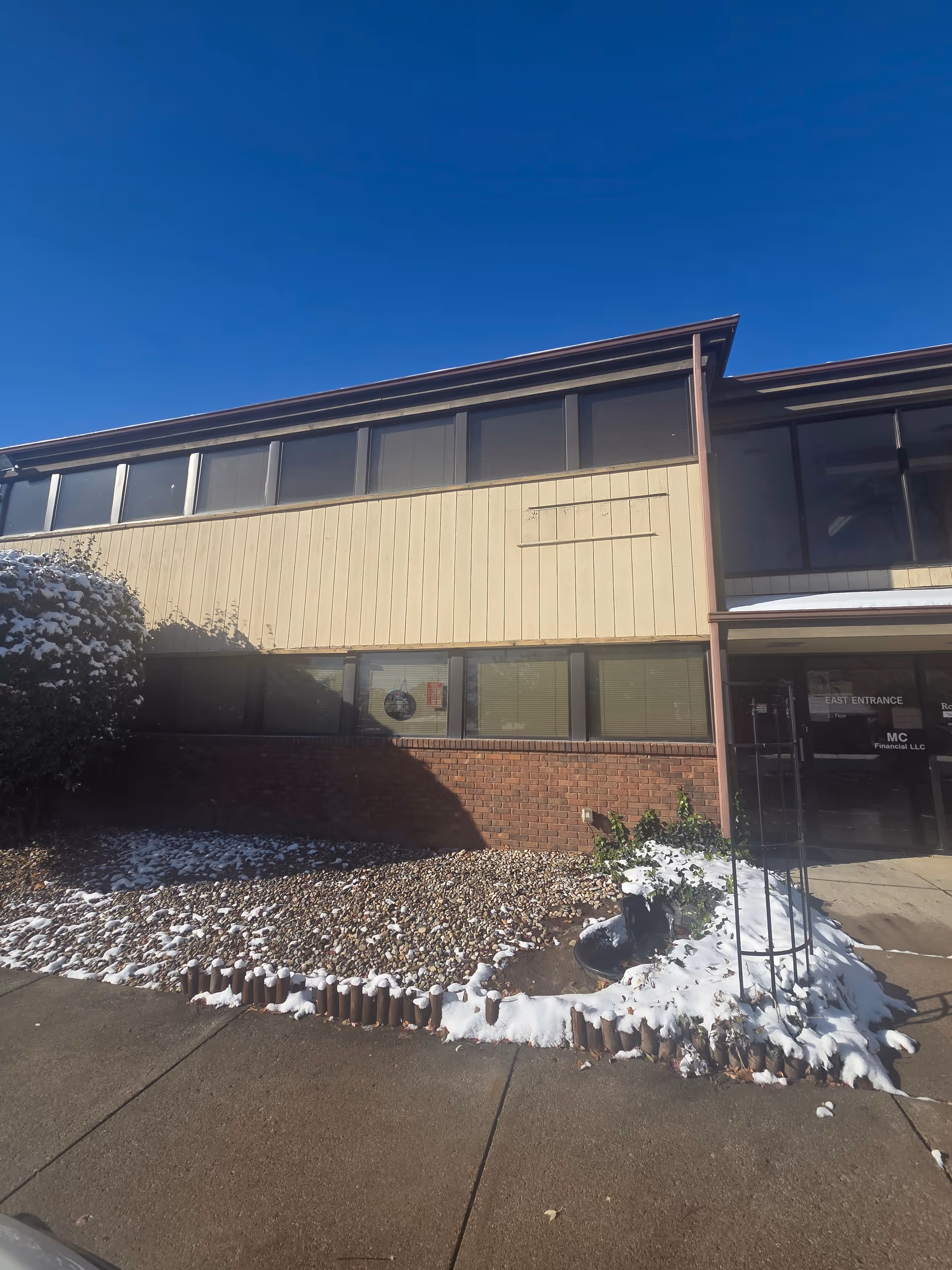Exterior view of a two-story building with beige siding and brick base, snow on landscaping.