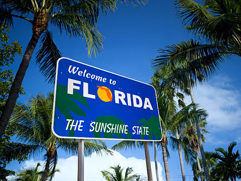 Blue Florida welcome sign surrounded by tall palm trees under a clear sky.