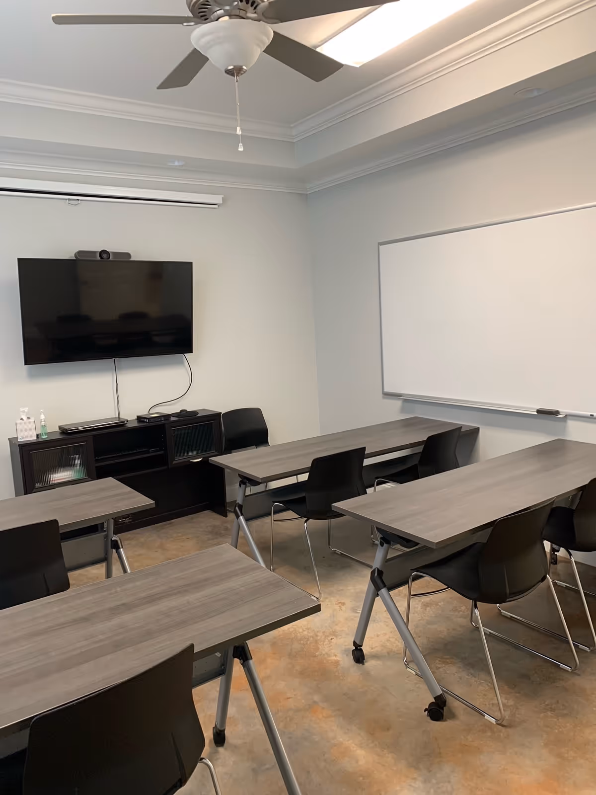 Classroom or training room with gray tables, black chairs, a wall-mounted TV, whiteboard, and ceiling fan.