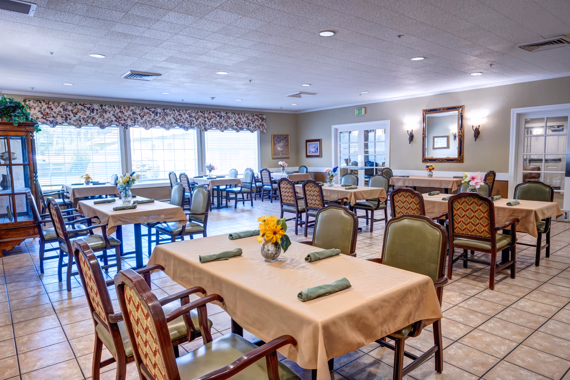 A bright dining room with multiple tables covered in beige tablecloths, each set with green napkins and small flower arrangements. The room has large windows with floral valances letting in natural light, tiled floors, and a mix of green and patterned chairs. There is a wooden cabinet on the left and a mirror with wall sconces on the right wall.