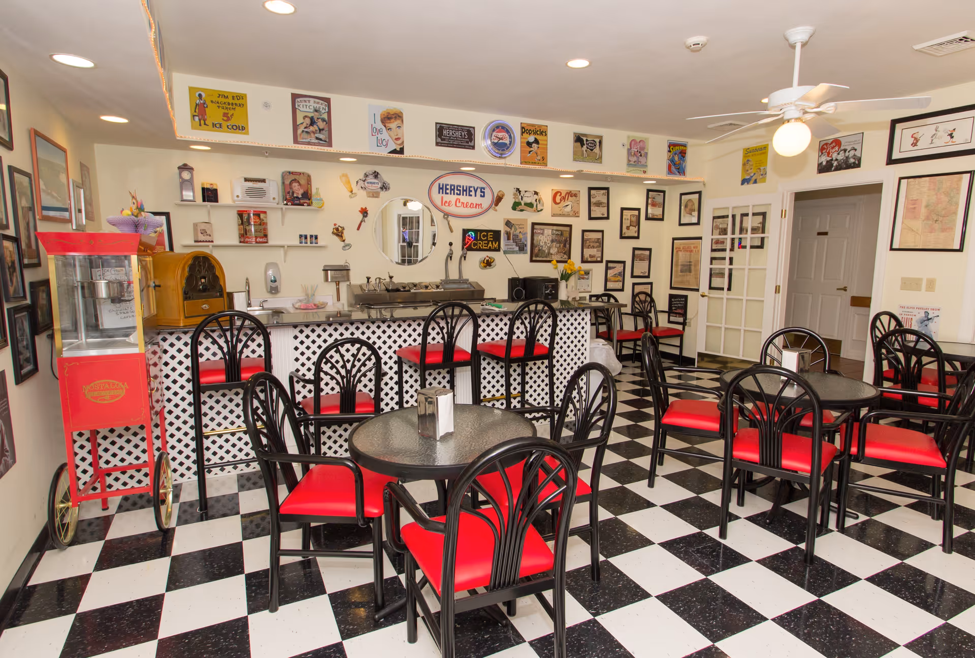 A retro-style dining area with black and white checkered floor tiles, black chairs with red cushions, and round tables. The back wall features a counter with a white lattice front, decorated with vintage signs and memorabilia, including a Hershey's Ice Cream sign. There is a red popcorn machine on the left side and a ceiling fan with a light on the right side.
