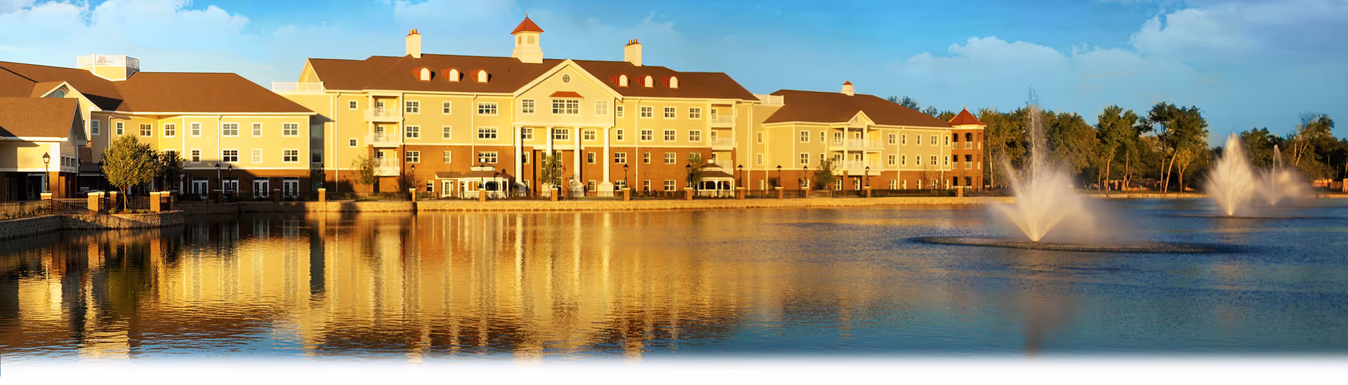 A large multi-story senior living building sits beside a lake with two water fountains and its reflection in the water.