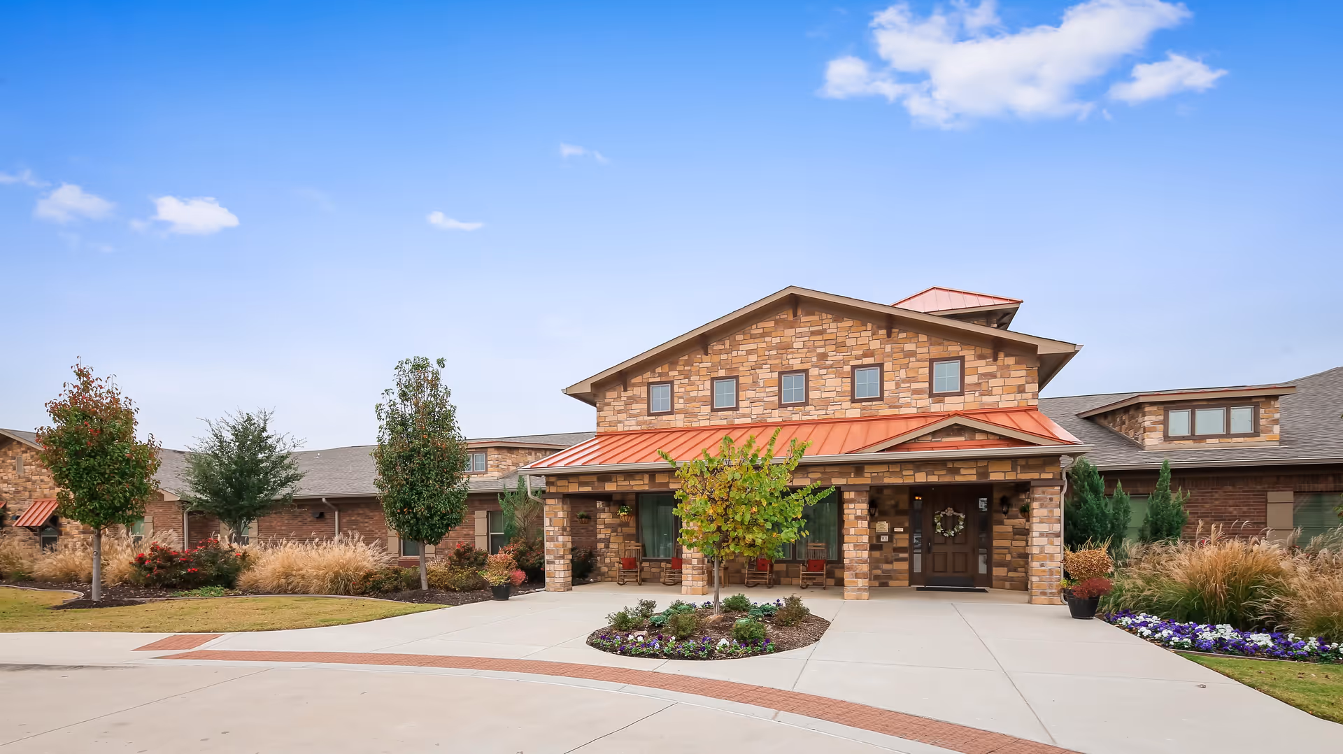 Front exterior view of Meadowood Assisted Living and Memory Care facility showing a large building with stone and brick facade, a red metal roof, several windows, a covered entrance with seating, landscaped garden beds, and a clear blue sky.