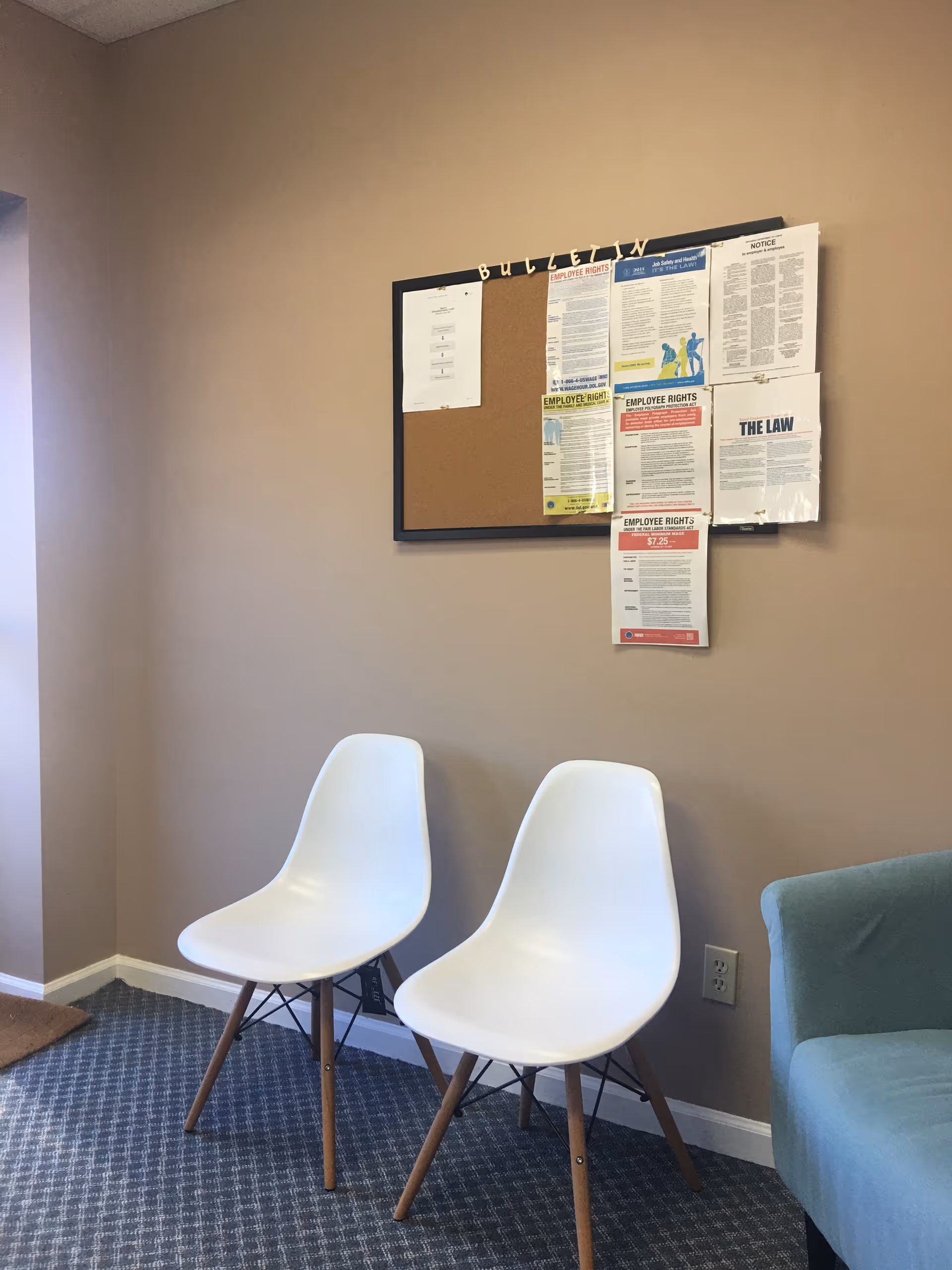 Two white chairs beside a bulletin board with scattered paperwork in a beige-walled waiting area.