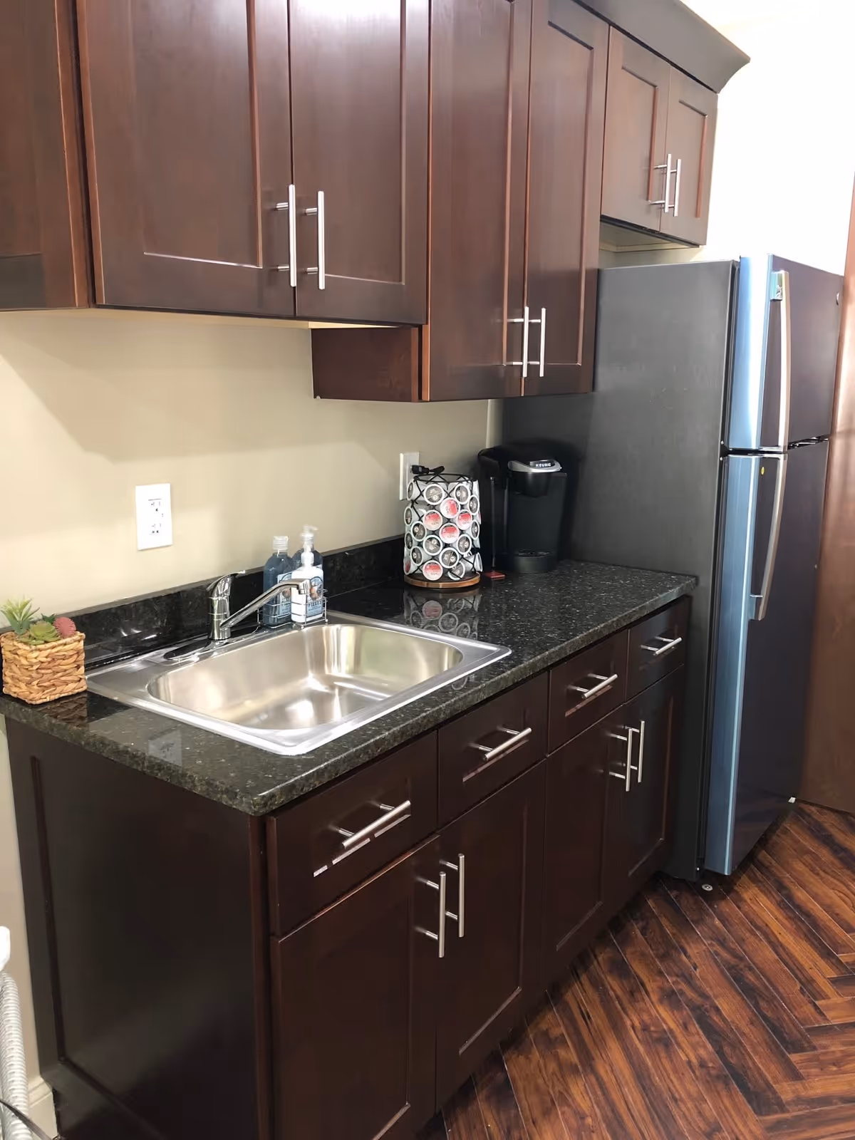 A kitchen area with dark wooden cabinets, a stainless steel sink, a black granite countertop, a coffee maker, and a refrigerator. The floor has a wood pattern.