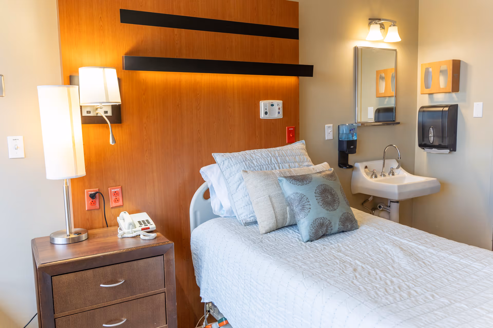 A neatly made single bed with multiple pillows in a room featuring a wooden headboard. Next to the bed is a wooden nightstand with a lamp and a telephone. On the wall beside the bed is a small sink with a mirror above it, a soap dispenser, and a paper towel dispenser.