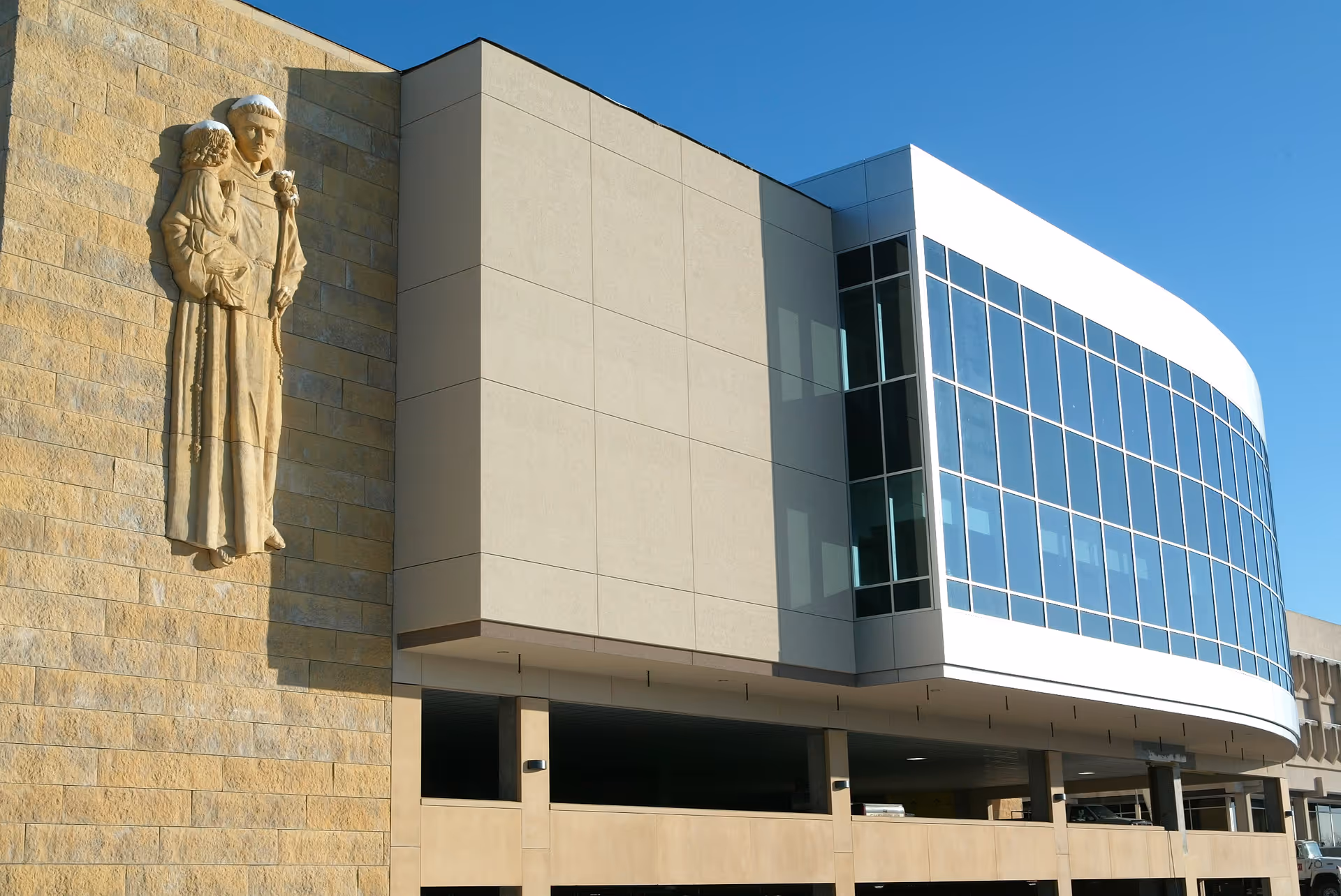Front exterior of a modern hospital with curved glass facade and a stone statue.
