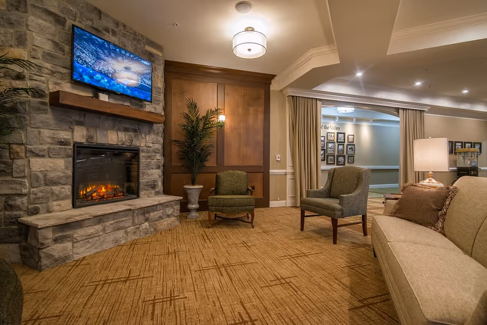 A cozy living room area in a senior living facility with a stone fireplace featuring a lit fire and a mounted flat-screen TV above it. The room has beige carpet with a geometric pattern, two green upholstered chairs, a beige sofa with brown pillows, a table lamp, and a potted plant against a wooden paneled wall. In the background, there is an open doorway with curtains and framed pictures on the wall.