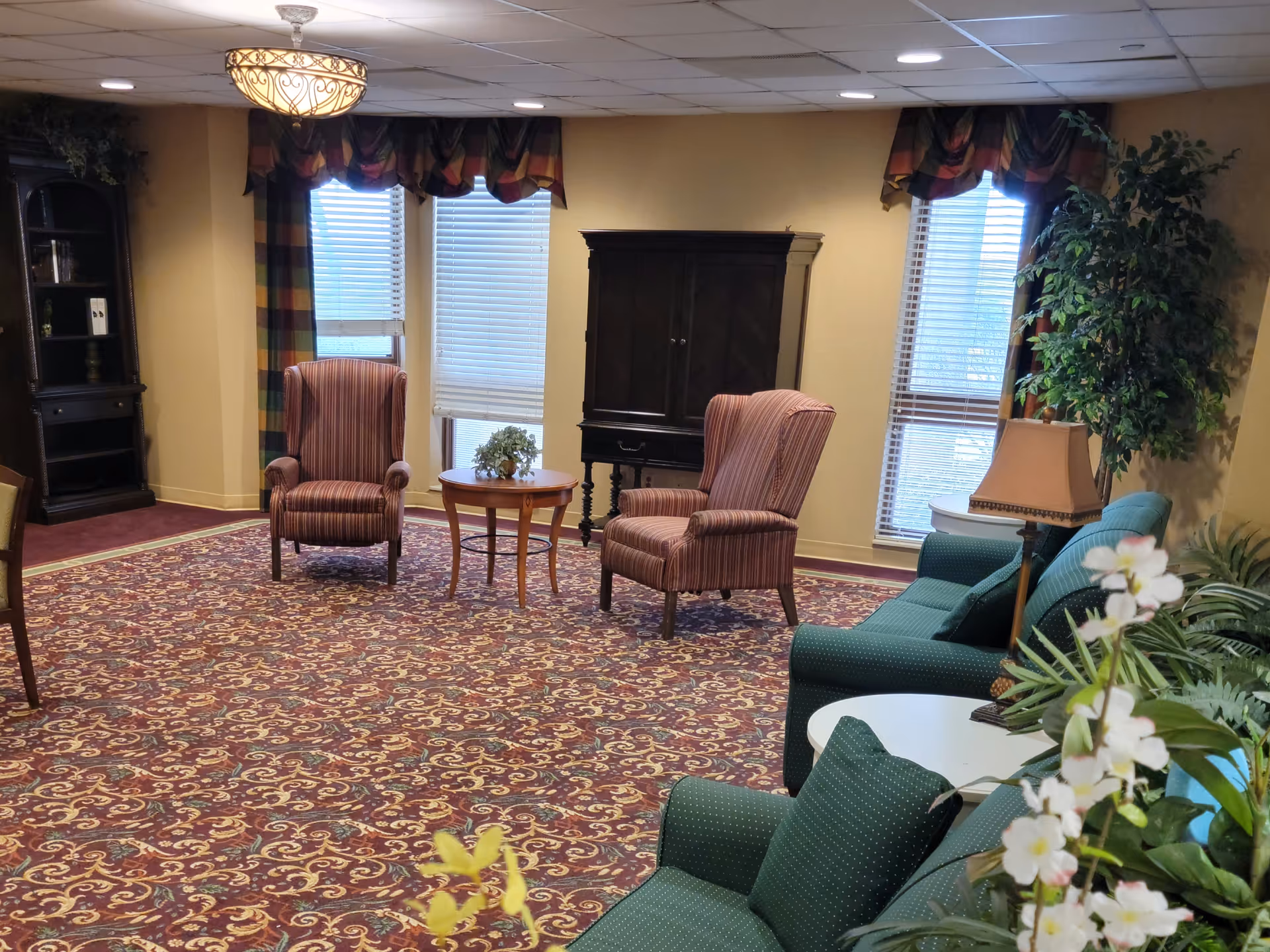 A cozy living room area with patterned carpet, two striped armchairs around a small round wooden table, a dark wooden cabinet against the wall, green upholstered sofas with cushions, a side table with a lamp, potted plants, and windows with blinds and colorful valances.