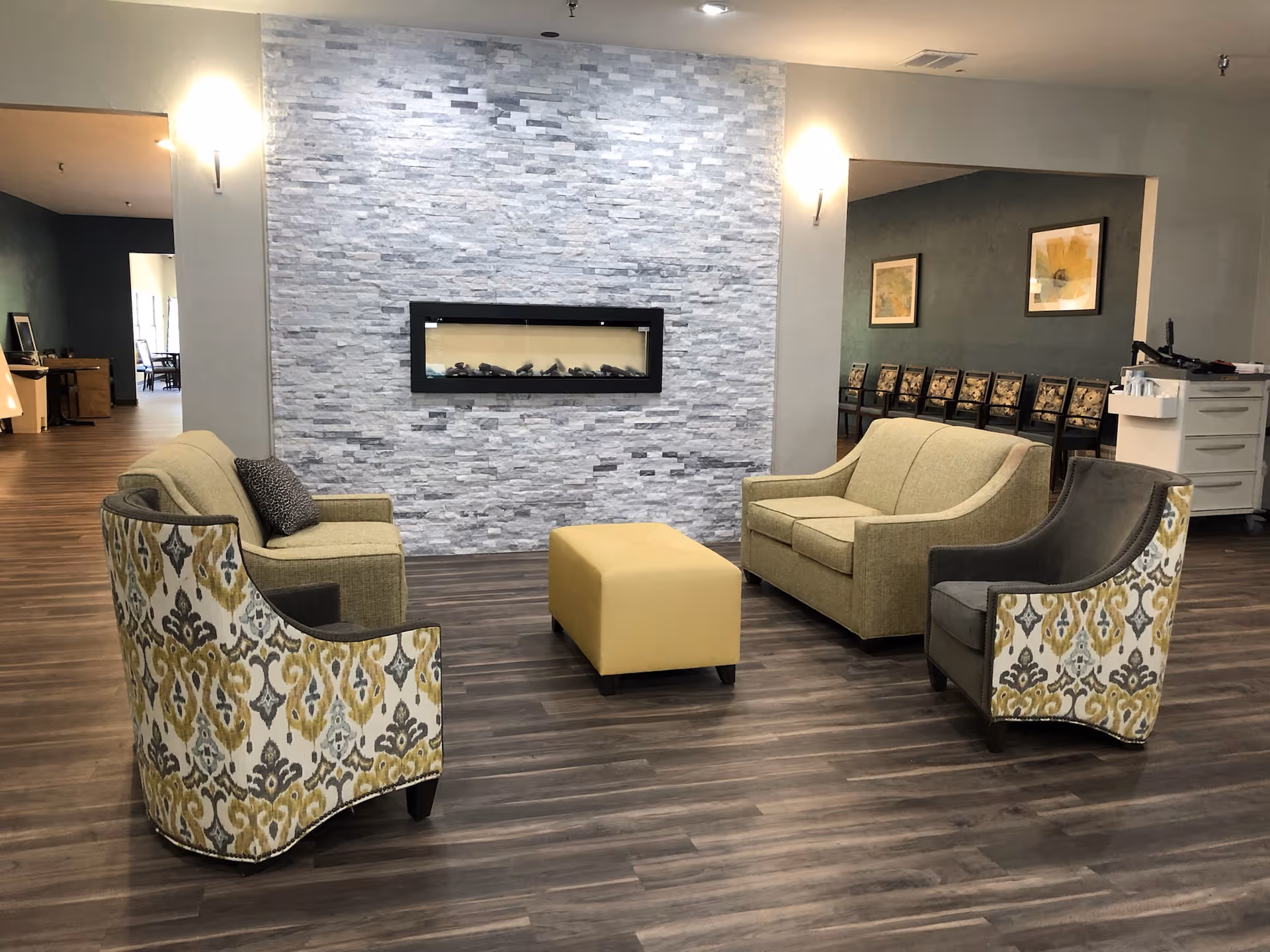 A modern seating area in a senior living facility with two patterned armchairs, a yellow ottoman, and two beige sofas arranged around a wall-mounted electric fireplace with a gray stone tile backdrop. The room has wood flooring and soft lighting, with additional chairs and framed artwork visible in the background.