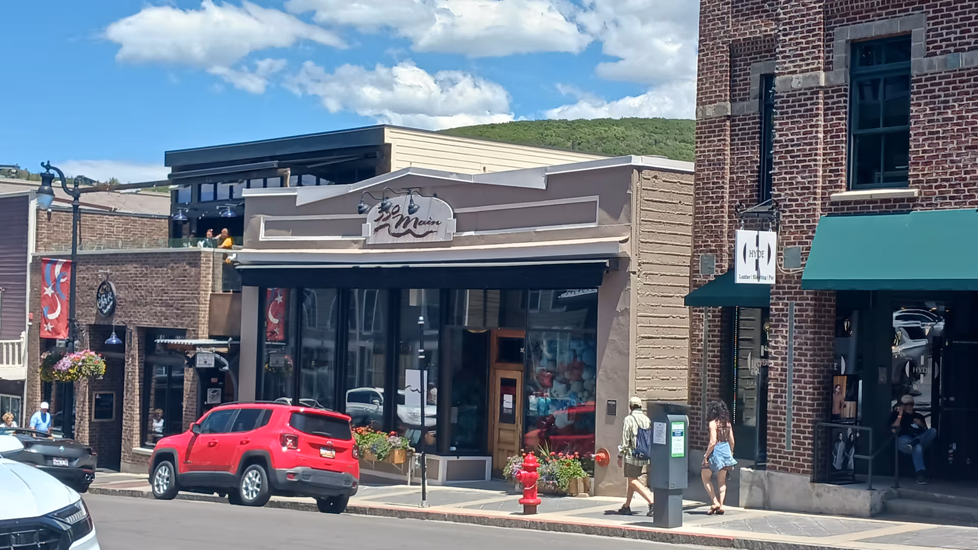 Downtown storefronts line a street with pedestrians and a red SUV parked nearby.