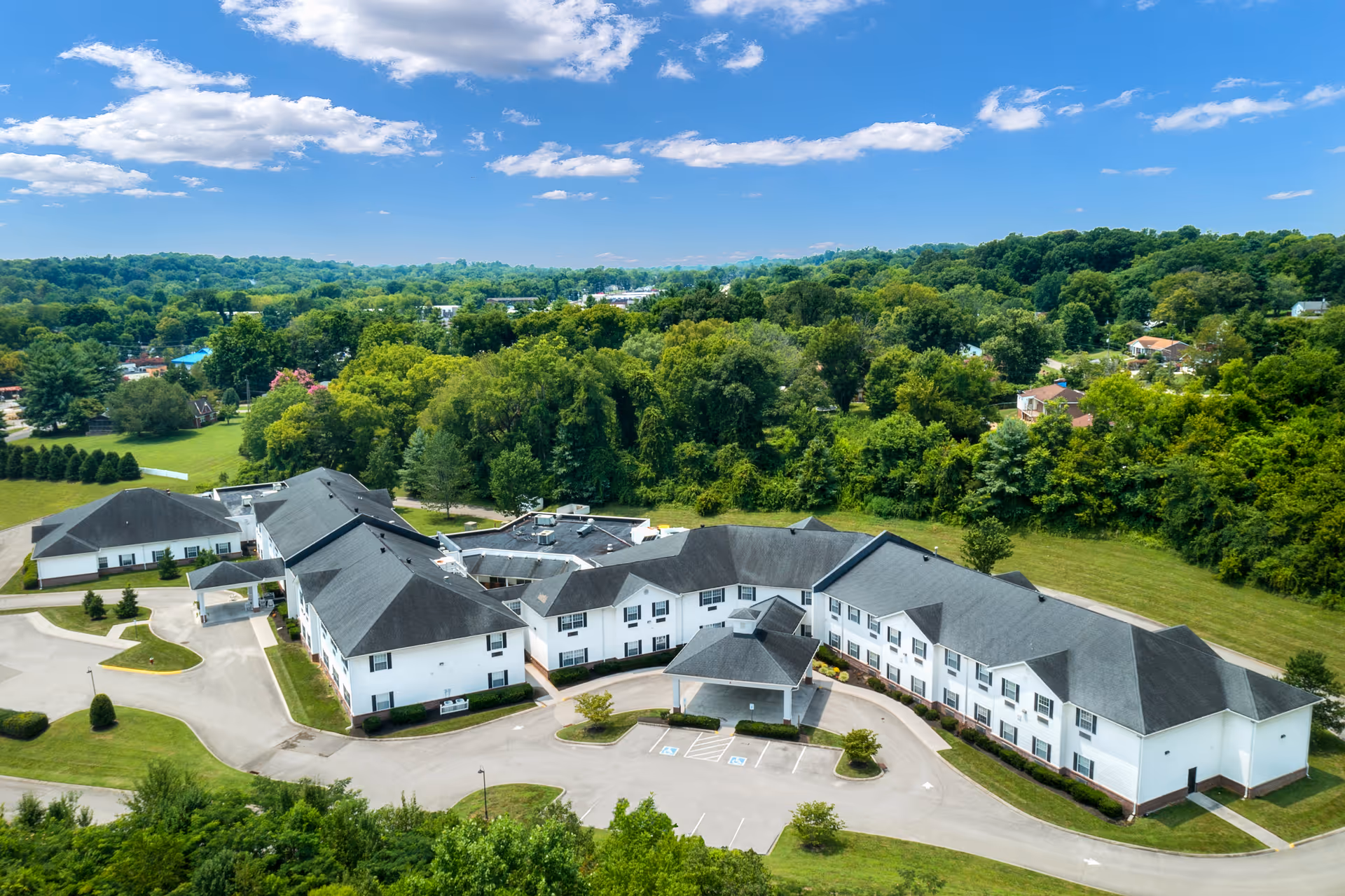 Aerial view of a large senior living facility named Trinity Hills of Knoxville surrounded by greenery and trees under a blue sky with some clouds. The building has a white exterior with a dark roof and a circular driveway with parking spaces in front.