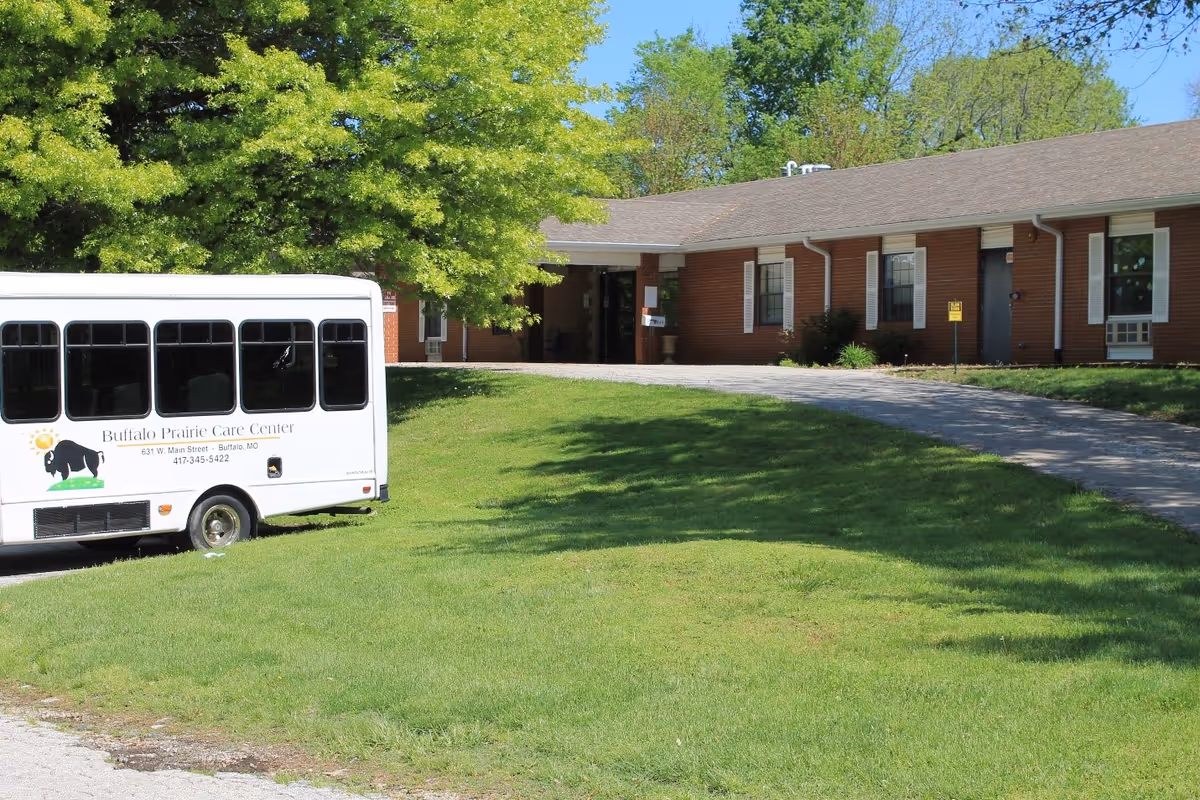 Front exterior of a single-story brick senior care facility with a Buffalo Prairie Care Center shuttle bus parked on the lawn and a large tree nearby.