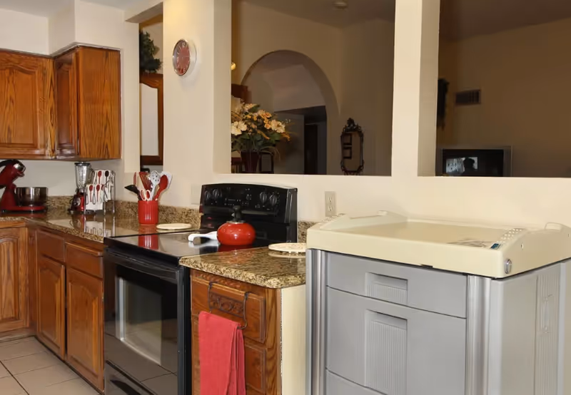 Kitchen in a senior facility featuring wooden cabinets, granite counters, stove, and a red kettle.