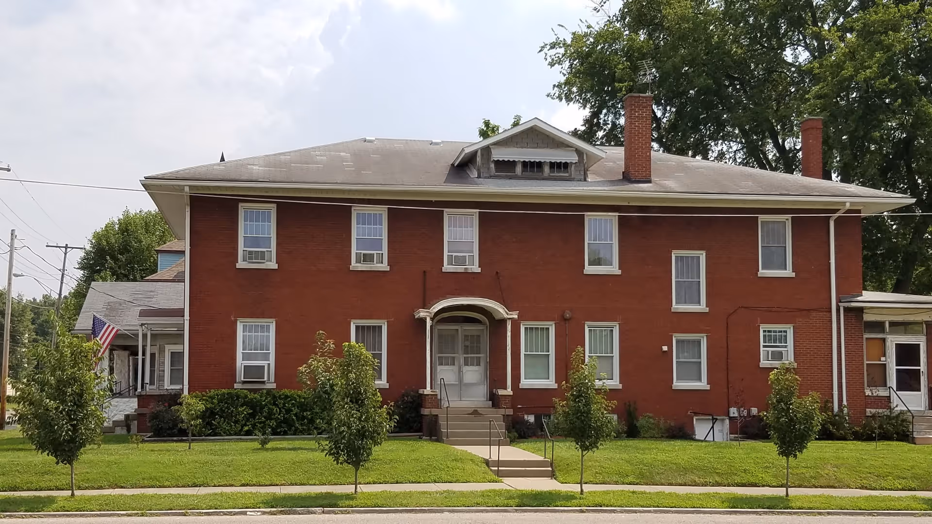 Front exterior view of a two-story red brick building with multiple windows, a central entrance with steps and a small porch, surrounded by a well-maintained lawn and small trees.
