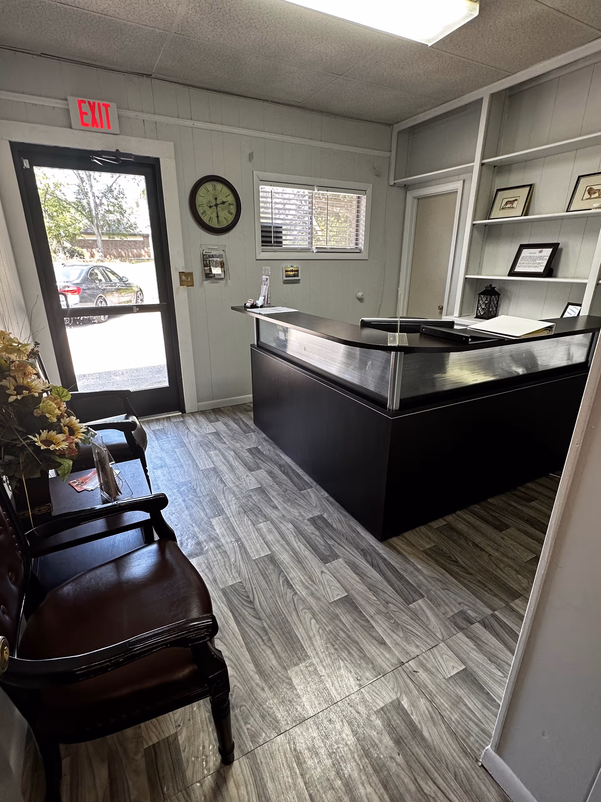 Reception area with a black-front desk, chairs, clock, shelves, and exit door.