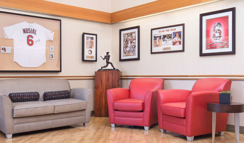 A cozy seating area with a gray couch and two red armchairs on a wooden floor. The walls are decorated with framed baseball memorabilia, including a white jersey with the name Musial and the number 6, several photos, and a bronze baseball player statue on a wooden pedestal.