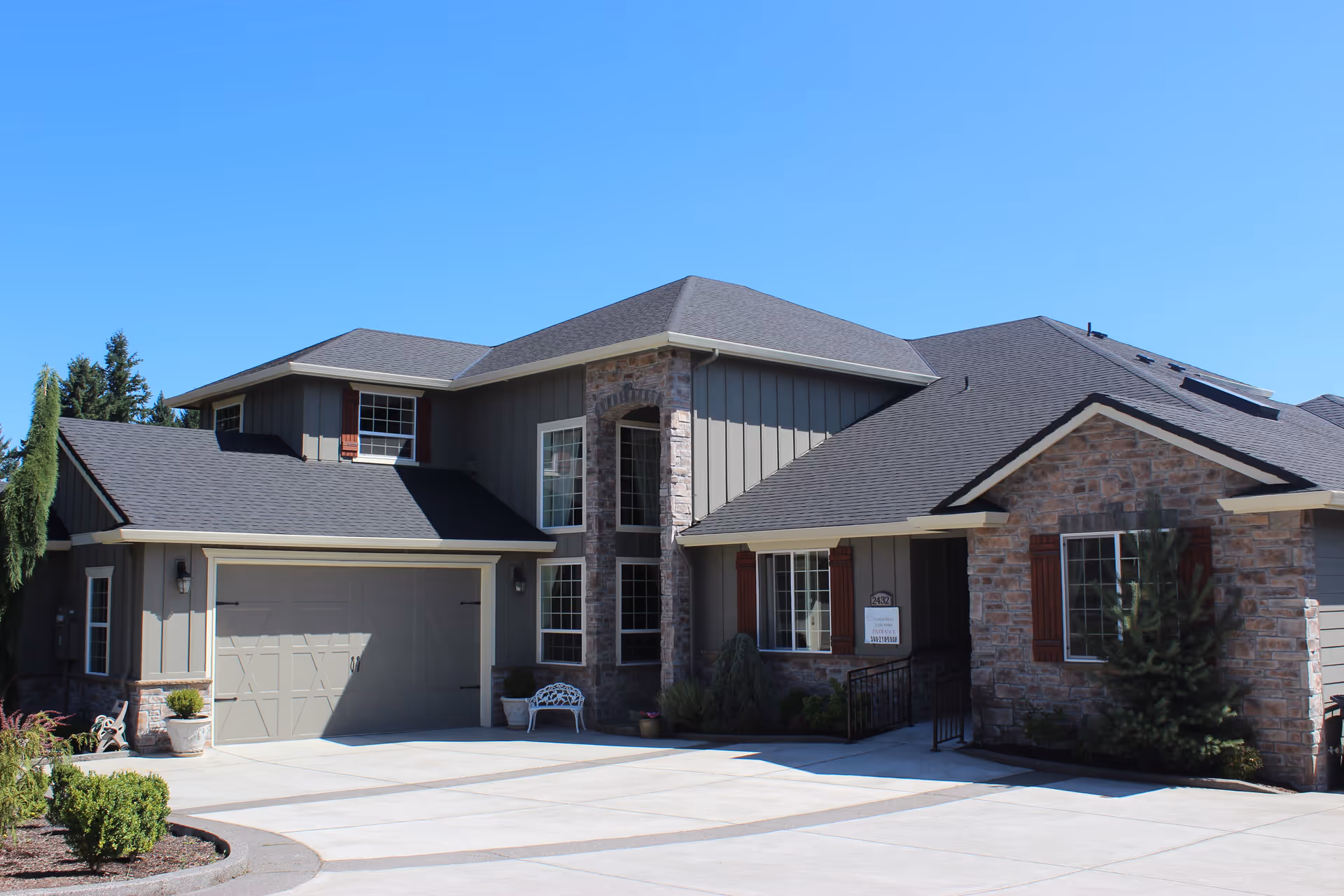 Exterior view of a modern two-story residential building with a combination of stone and wood siding, a large driveway, and a garage under a clear blue sky.
