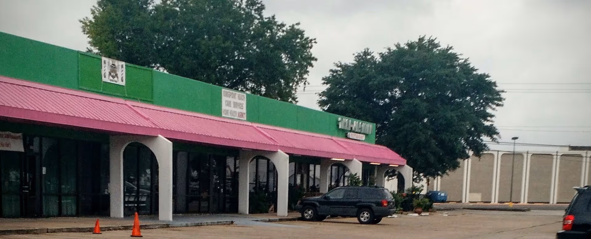 Row of storefronts with green facade and pink awning along a parking lot.