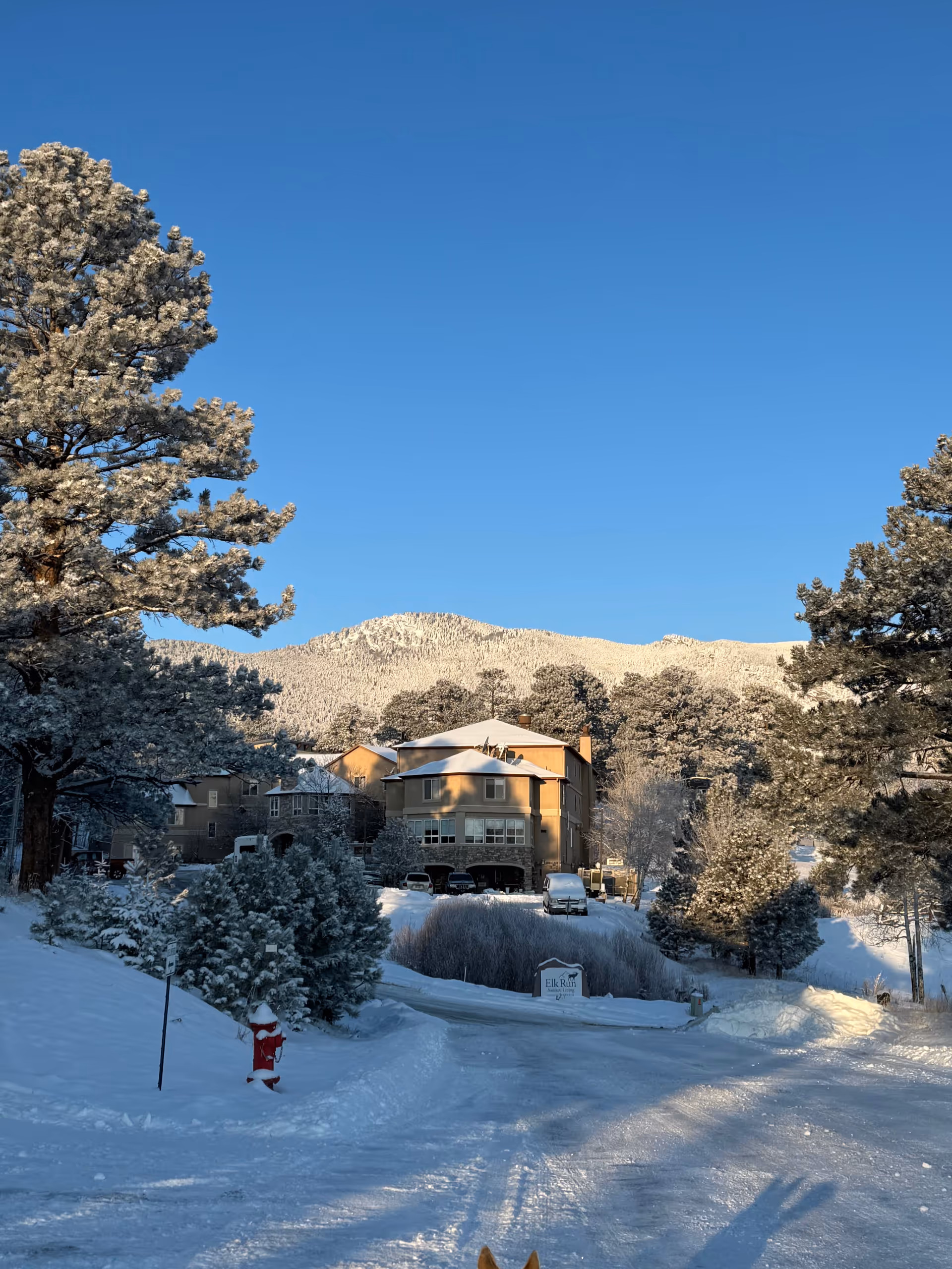 Snow-covered Elk Run Assisted Living building with trees and mountain backdrop.