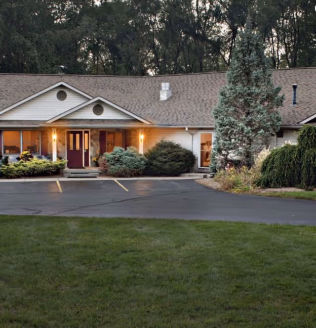 Exterior view of a single-story senior living facility building with a gable roof, front entrance with double doors, lit porch lights, surrounding bushes, trees, and a paved parking area in front.
