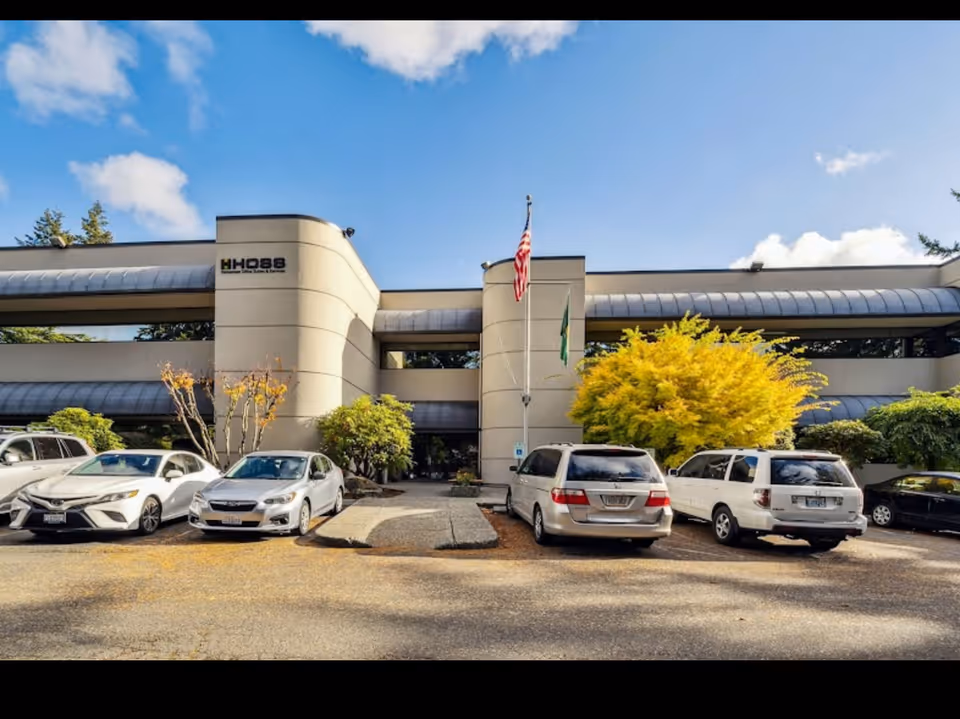 Modern senior care facility exterior with parked cars and flagpoles in front.