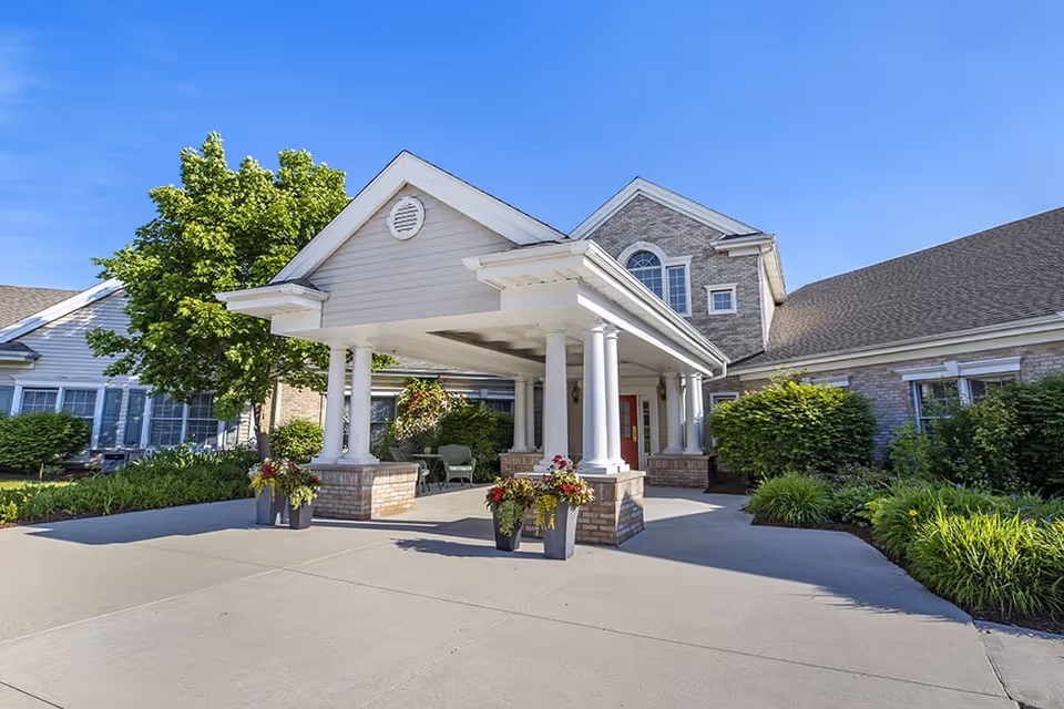 Front entrance of Brookdale Troy featuring a covered porte-cochere with white columns, planters, and landscaped grounds under a clear blue sky.