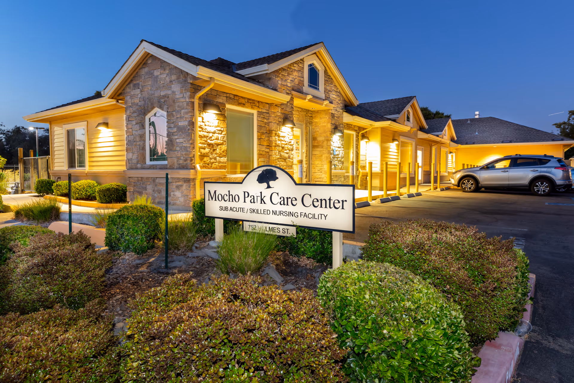 Exterior view of a stone-clad senior care facility entrance with landscaping and sign.