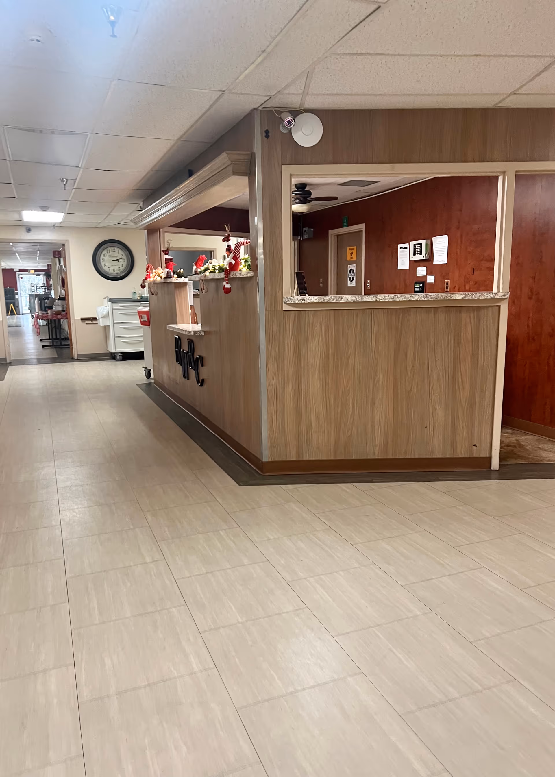 Interior view of a nursing facility hallway with a reception desk decorated with holiday ornaments. The floor is tiled, and there is a clock on the wall. The reception area has wood paneling and a granite countertop.