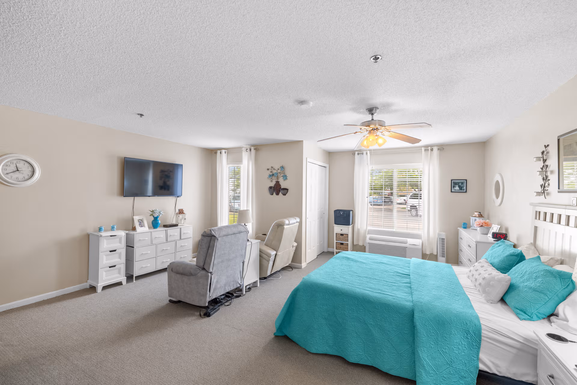 Bright bedroom suite with teal bedding, two recliners, white dressers, and a wall-mounted TV.