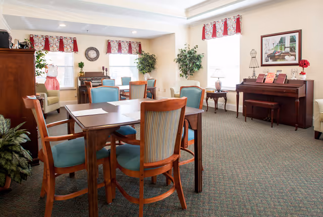 A bright and cozy common area in a senior living facility featuring wooden tables and chairs with blue cushions, a piano against the wall, decorative plants, framed artwork, and windows with red and white valances allowing natural light to fill the room.