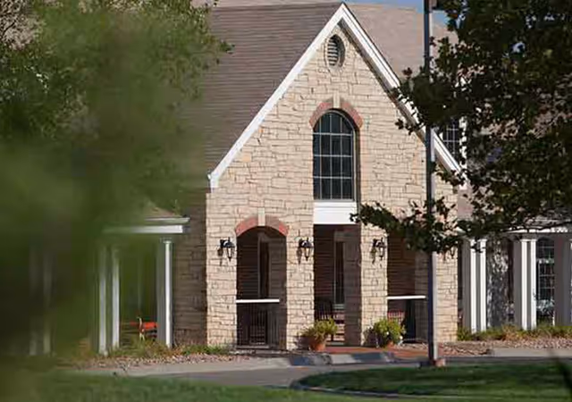 Front exterior of a stone-faced building with arched porch openings, a tall arched window, potted plants, and surrounding trees.