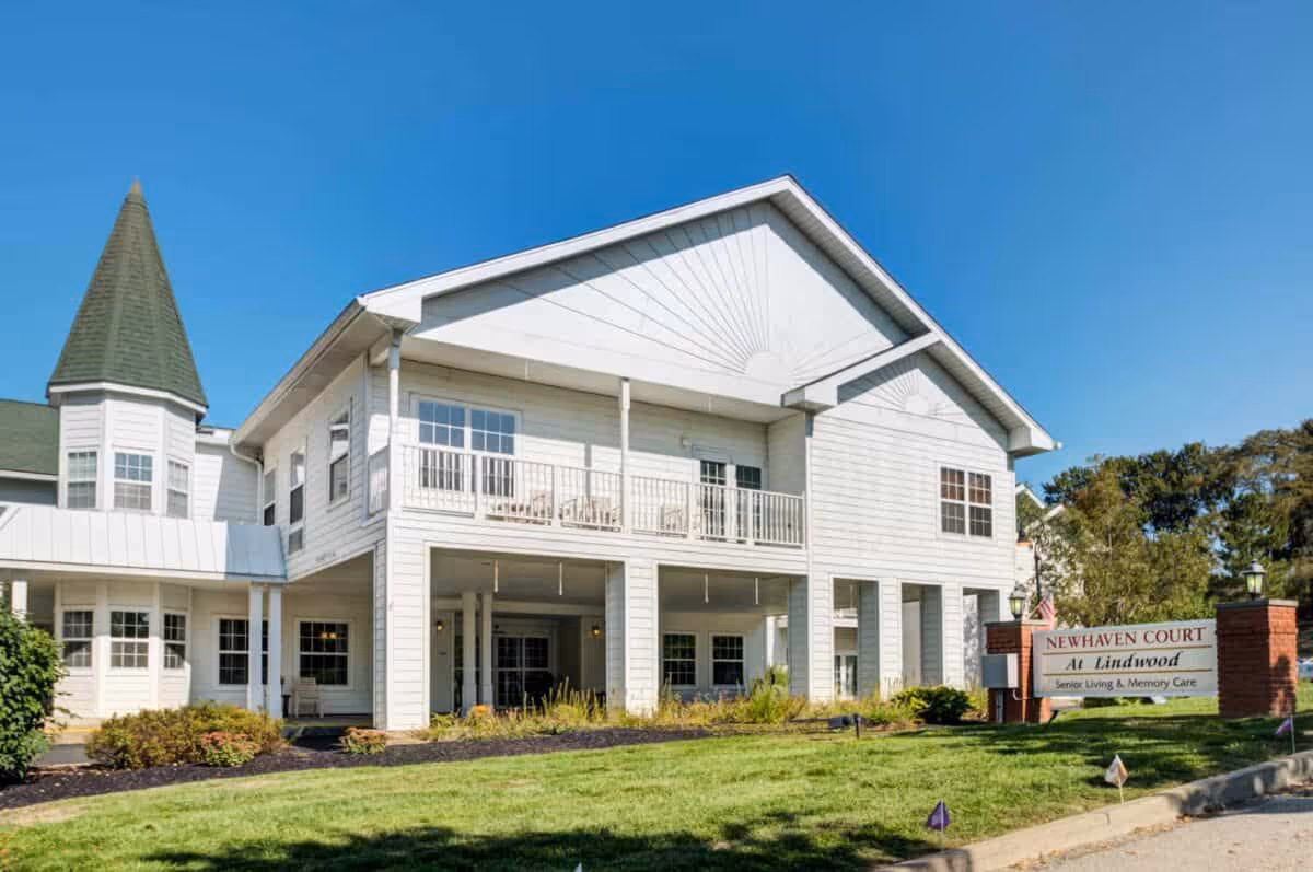 Exterior view of Newhaven Court at Lindwood, a senior living and memory care facility. The building is white with a green roof and features a covered entrance with columns, a balcony with chairs, and a turret on the left side. There is a well-maintained lawn and landscaping in front of the building, along with a sign that reads 'Newhaven Court at Lindwood Senior Living & Memory Care'. The sky is clear and blue.